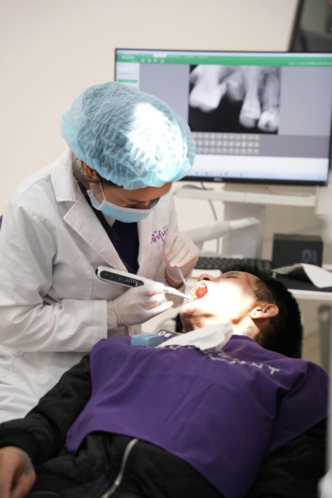 Dentist examines patient in clinic with medical equipment and X-ray display.